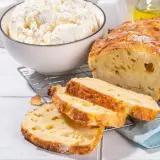 Chleb z twarogu, świeżo upieczony, z chrupiącą skórką i puszystym wnętrzem. Obok miska pełna białego twarogu, składnika tego pysznego wypieku.