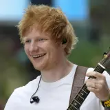 Ed Sheeran gra na gitarze, uśmiecha się szeroko. Fani w Polsce czekają na jego koncert.