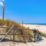 Rower czeka przy płocie na plaży, gotowy do odkrywania tras rowerowych nad morzem. W tle błękitne niebo i morze.