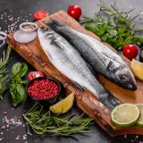 Dwie świeże ryby bass na desce, otoczone cytryną, limonką, pomidorkami, cebulą, rozmarynem i pieprzem.