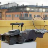 En abstrakt skulptur i mörk metall, placerad på ett vitt podium. I bakgrunden syns byggnader och en gul vägg, kanske vid museum Vasaparken.