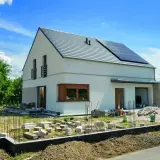 Nowy dom kostka z białym tynkiem i kamienną elewacją, na dachu panele słoneczne. Wokół plac budowy.