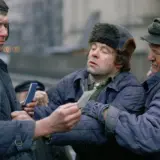 Trzech mężczyzn w zimowych kurtkach i czapkach, w tym jedna z futrzanymi uszami, dyskutuje, trzymając w rękach dokumenty. Scena z filmu "Miś" z niezapomnianą obsadą.