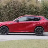 Czerwona Mazda CX-60 3.3 diesel z czarnymi felgami stoi na żwirowej drodze, za nią gęsty żywopłot.