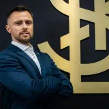 Mężczyzna w garniturze, trener ŁKS Łódź, stoi przed złotym logo klubu.