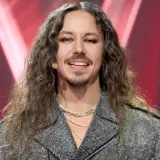 Michał Szpak w błyszczącej kurtce i z długimi włosami, uśmiecha się do kamery.