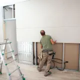 Pracownik wykonuje montaż płyt gipsowych na wkręty do stelaża. Obok stoi drabina.