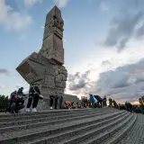 Uroczyste zwiedzanie Westerplatte. Monument, żołnierze w mundurach, wieńce i chmury na niebie tworzą podniosłą atmosferę.