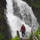 Mujer contemplando una cascada en Vielha e Mijaran. El agua cae con fuerza sobre rocas oscuras, rodeada de vegetación verde.