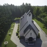 Kościół otoczony lasem, z wieżą i dzwonami. Wokół zadbane alejki i młode drzewka, tworzące urokliwe przypki.