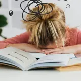 Dziewczynka z blond włosami w koku, zmęczona nauką, opiera głowę na otwartych książkach. Wokół jej głowy rysunki symbolizujące chaos i zmęczenie.