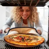 Uśmiechnięta kobieta wyjmuje z piekarnika pyszną pizzę. Widać, na jakim programie piec pizze, bo jest idealnie wypieczona.