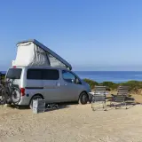 Silberner Camper mit Dachzelt und Fahrrädern am Heck, zwei Liegestühle und ein Tisch stehen bereit für entspanntes italien wildcampen am Meer.
