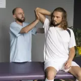 Fizjoterapeuta bada naciągnięty bark młodego sportowca siedzącego na kozetce.