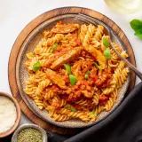 Pyszny makaron z kurczakiem w sosie pomidorowym, udekorowany bazylią. Obok miseczki z parmezanem i ziołami.