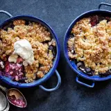 Två blåa ugnsformar med glutenfri smulpaj fylld med bär och toppad med grädde. Serveras med antika skedar.