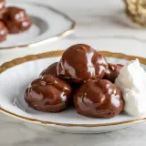 Profitterol ricetta: golosi bignè ricoperti di cioccolato fondente, serviti con panna montata su un piatto bianco con bordo dorato.
