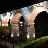 Nocna iluminacja kamiennego wiaduktu z ceglanymi łukami. Widać fragment mostku i drzewa. To piękna bytów trasa.