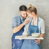 Para uczciła 9 rocznicę ślubu, tworząc ceramikę. Mężczyzna delikatnie dotyka nosa kobiety, oboje ubrani w fartuchy, z gliną na twarzach.
