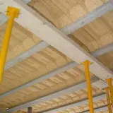 Strop rector w budowie, podparty żółtymi podporami. Widoczne belki drewniane i płyty stropowe.