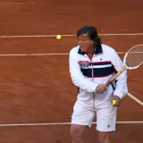 Panatta w białej kurtce i spodenkach, z rakietą i piłką tenisową na korcie.