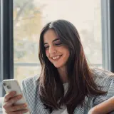 Uśmiechnięta kobieta trzyma telefon, czeka na wiadomość od ukochanej osoby. To piękny przykład, jak technologia wspiera związek na odległość.