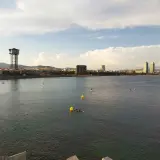 Vista de la playa y el mar con el icónico hotel vela al fondo. Personas disfrutan del agua en tablas de paddle surf.