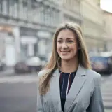 Uśmiechnięta kobieta w marynarce na tle miasta. Czytała pozytywne renee opinie forum i postanowiła wybrać się na spacer.
