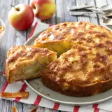 Un délicieux gâteau aux pommes, doré et moelleux, avec des morceaux de fruits visibles. Des pommes fraîches et des chips de pommes complètent la scène.