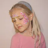 Ein Mädchen mit blonden Haaren und einem Stirnband, das ein Blumenmuster auf der Wange trägt. Das Stern schminken Kinder ist hier als zarte Blüten umgesetzt.