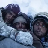 Tre uomini stremati nel gelo, un'immagine che evoca le recensioni de "La società della neve", la lotta per la sopravvivenza.