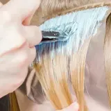 Dłoń z pędzlem nakłada jasnoniebieski rozjaśniacz na pasma blond włosów. Czas ekspozycji jest kluczowy, by uzyskać pożądany efekt.