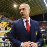 Sylvinho w granatowym garniturze i czerwonym krawacie, z emblematem na piersi, stoi na tle trybun stadionu.