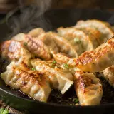 Gorące, rumiane pierożki gyoza parują na patelni. Jedna sztuka jest podnoszona pałeczkami, gotowa do zanurzenia w sosie.