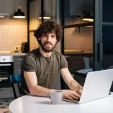 Uśmiechnięty mężczyzna z kręconymi włosami pracuje na laptopie w kuchni, planując, jak zapłacić czynsz.