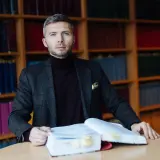 Mężczyzna w garniturze siedzi przy stole z otwartą księgą, otoczony regałami pełnymi książek. To jego zabezpieczenie STB.