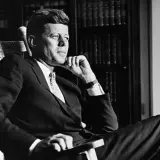 John F. Kennedy sitzt nachdenklich in einem Schaukelstuhl, seine Hand am Kinn. Trotz seiner bekannten **John F. Kennedy Krankheit** strahlt er eine ruhige Entschlossenheit aus.