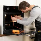 Technik sprawdza, czy piekarnik elektryczny ma bezpiecznik, używając multimetru. Obok stoi skrzynka z narzędziami.