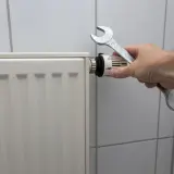 Dłoń z kluczem nastawia termostat, pokazując, jak spuścić wodę z kaloryfera.