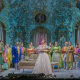 Primadonna w lśniącej sukni śpiewa na scenie operowej, otoczona przez barwnie ubranych artystów.
