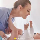 Uśmiechnięta mama pochyla się nad dzieckiem, które leży na plecach. Wykonuje szerokie pieluchowanie, delikatnie unosząc nóżki malucha.