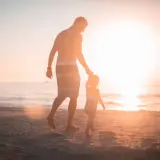 Ojciec prowadzi za rękę swoje dziecko 4 lata, spacerując po plaży o zachodzie słońca.