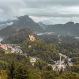 Schlechtes Wetter trübt den Urlaub. Ein gelbes Schloss thront auf einem bewaldeten Hügel über einem Dorf am See.