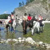 Konni jeźdźcy na koniach w rzece, w tle skaliste zbocze i plaża z ludźmi. To widok z Bukowiny Tatrzańskiej, jakby rynek atrakcji.