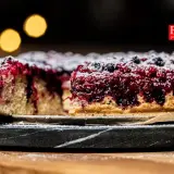 Pyszne ciasto z porzeczkami, posypane cukrem pudrem, na ciemnym talerzu. W tle rozmyte, ciepłe światła.