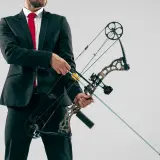 Mężczyzna w garniturze i czerwonym krawacie trzyma łuk rambo z zamontowaną strzałą.