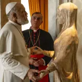 Papież w białej sutannie i kardynał w czerni rozmawiają przy kamiennej rzeźbie. Scena z serialu "nowy papież serial".