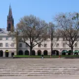 Łódź rynek z kamienicami, drzewami i wieżą kościelną w tle.