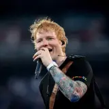 Ed Sheeran śpiewa do mikrofonu, ma tatuaże na rękach i słuchawki.