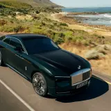 Elegancki, ciemnozielony Rolls-Royce elektryczny sunie po malowniczej drodze nad oceanem.